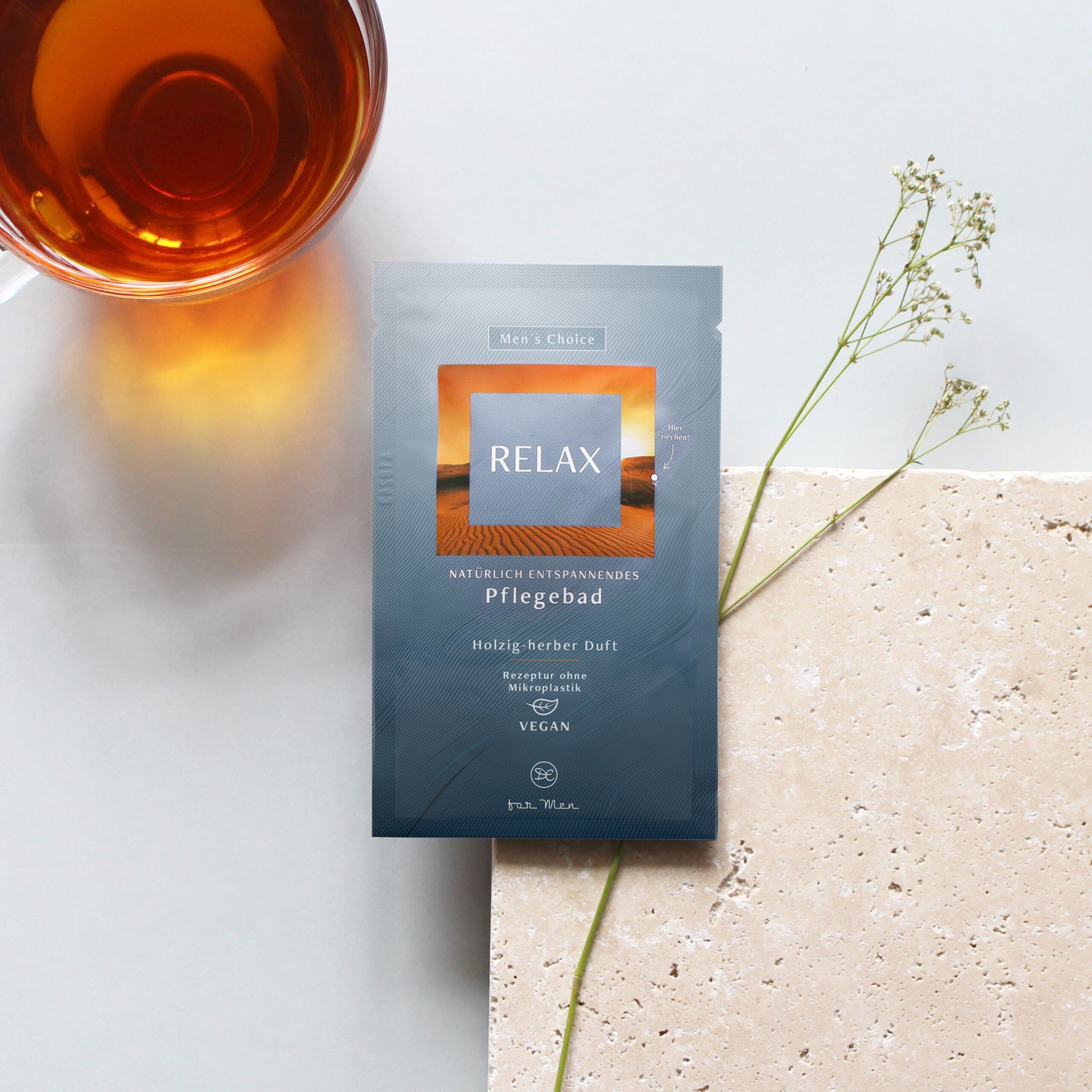 Enkele verpakking RELAX op licht oppervlak. Naast verpakking: glas met vloeistof, plant. Tekst: Verzorgend bad, houtachtig-kruidige geur, vegan.