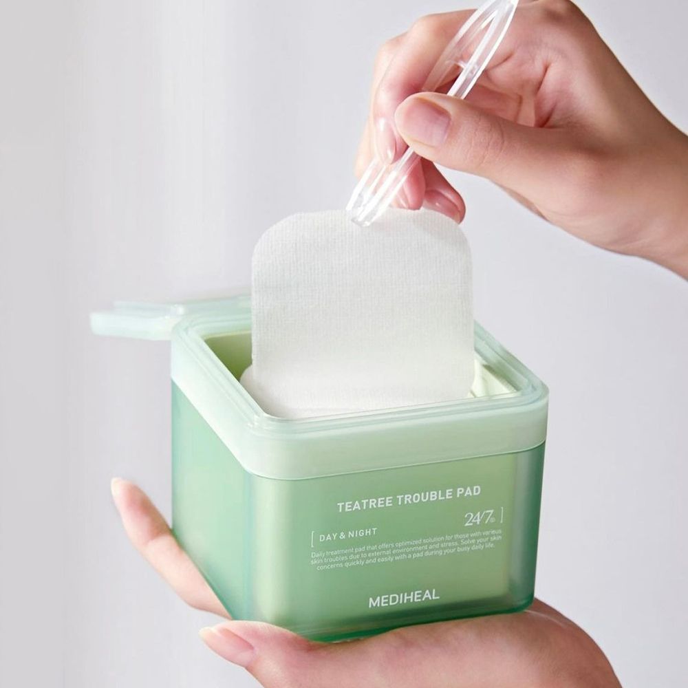 Boîte verte avec tampons. Une main tient la boîte, l'autre retire un tampon avec une pince. Inscription : TEA TREE TROUBLE PAD.