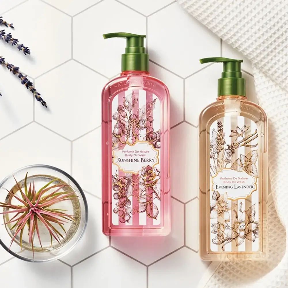 Twee flessen Body Oil Wash met bloemmotieven. Eén roze, één beige. Groene pompen. Opschriften Sunshine Berry en Evening Lavender.