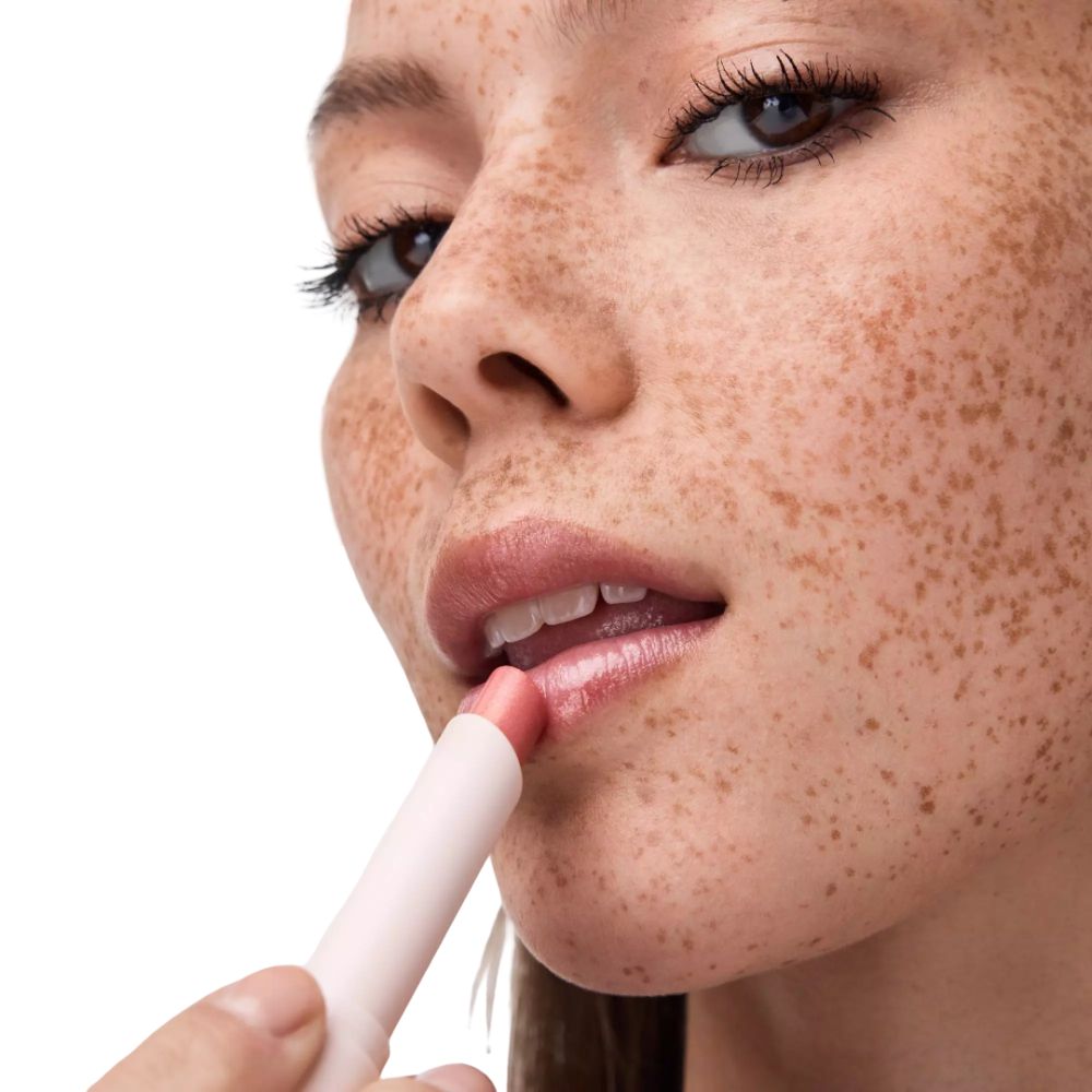 Close-up van een vrouw die lipgloss aanbrengt. Roze lipstick, lichte huid, sproeten.