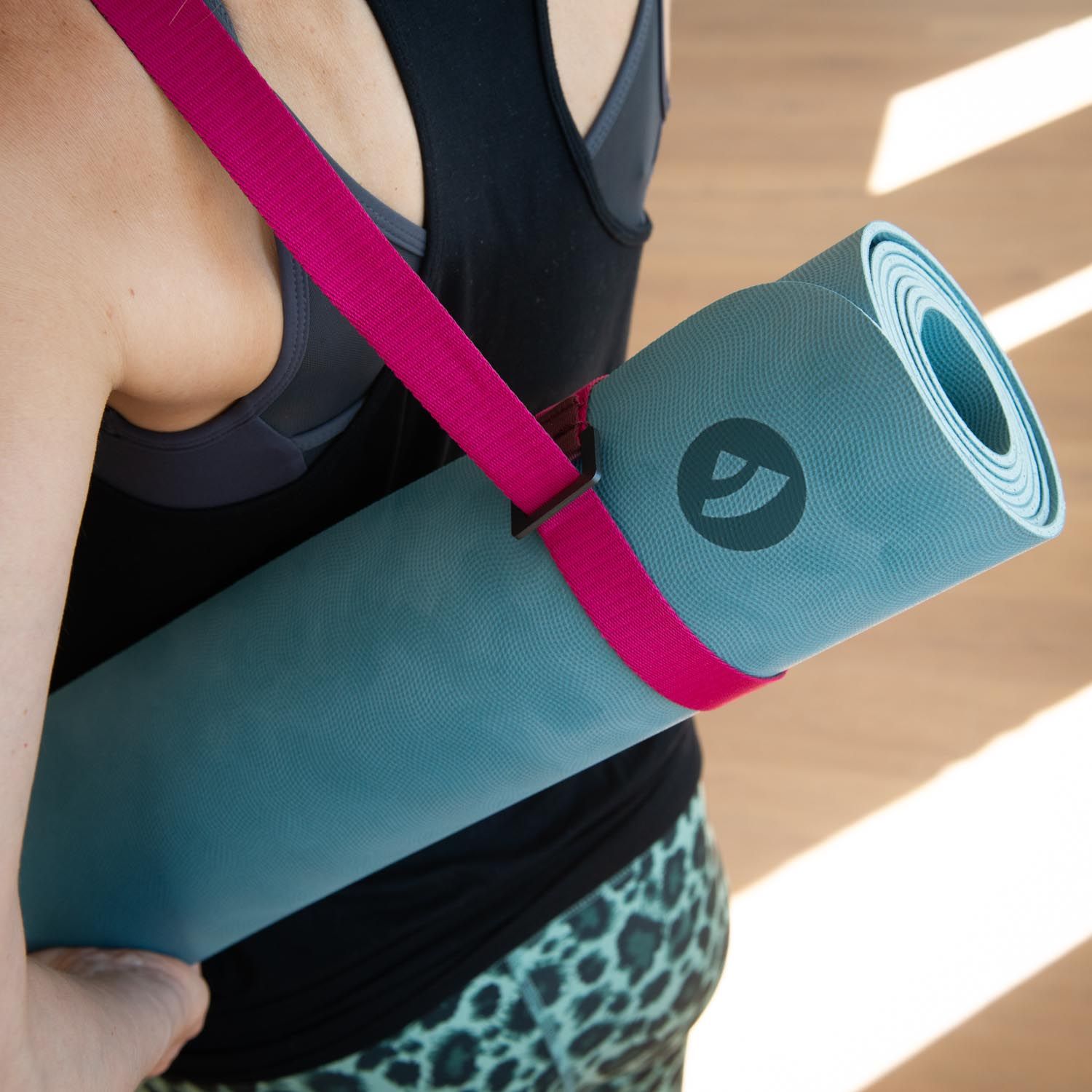 Persoon draagt opgerolde yogamat met roze draagriem. Logo op de mat. Persoon van achteren gezien.