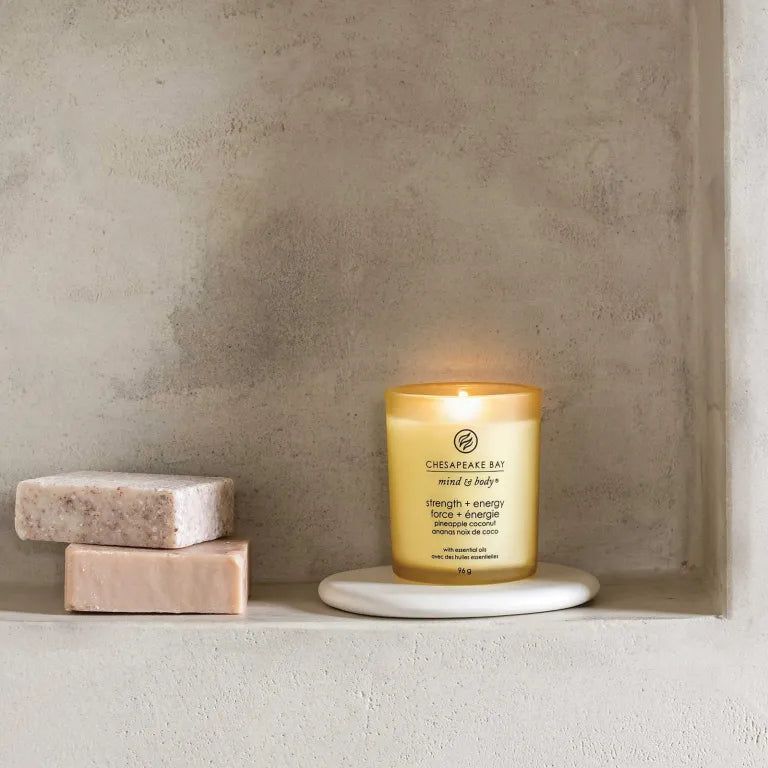 Bougie jaune Chesapeake Bay mind & body, force + énergie, ananas noix de coco, sur socle blanc. Deux savons à côté.