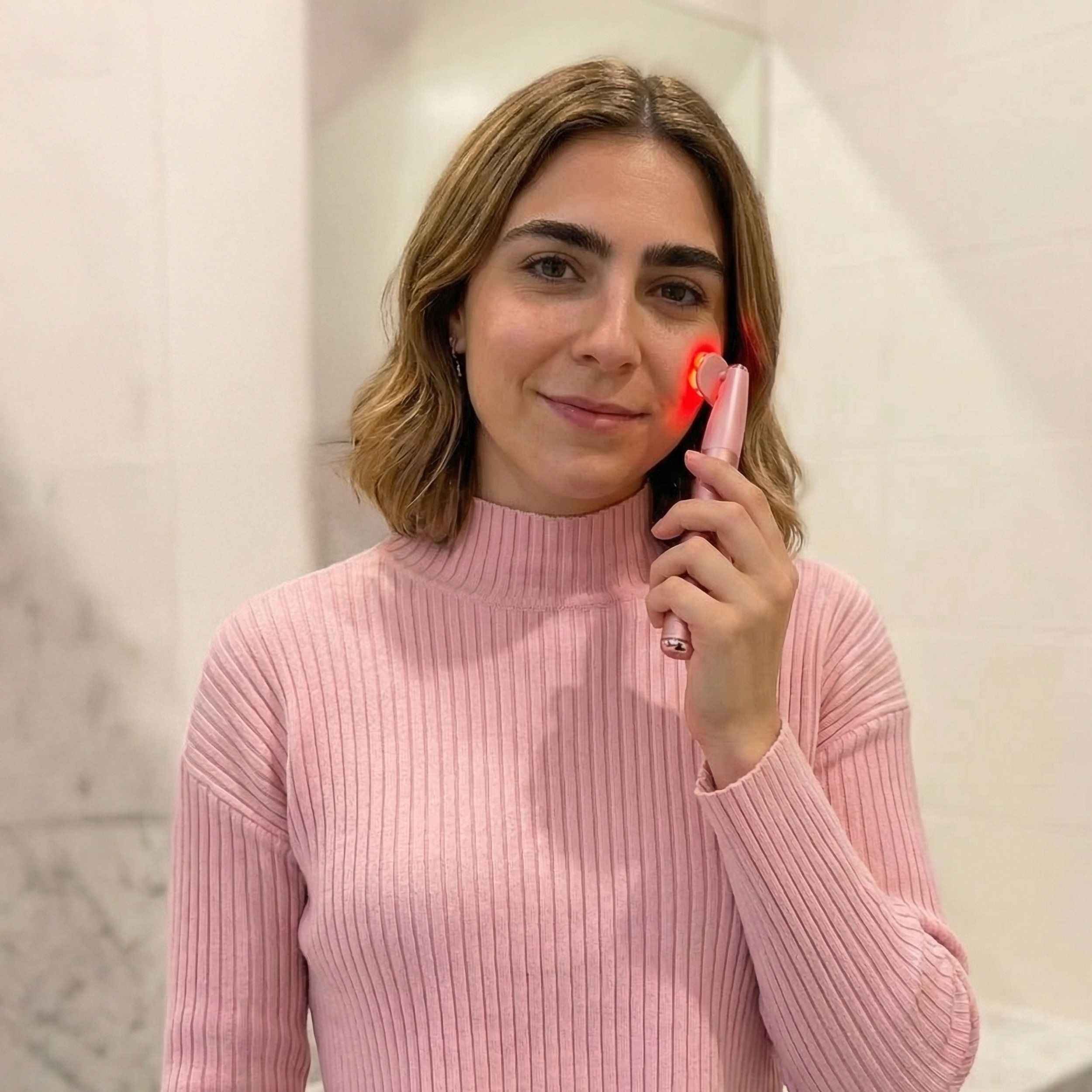 Personne tenant un appareil de soin rose. Lumière rouge sur la peau. L'appareil a une poignée allongée.