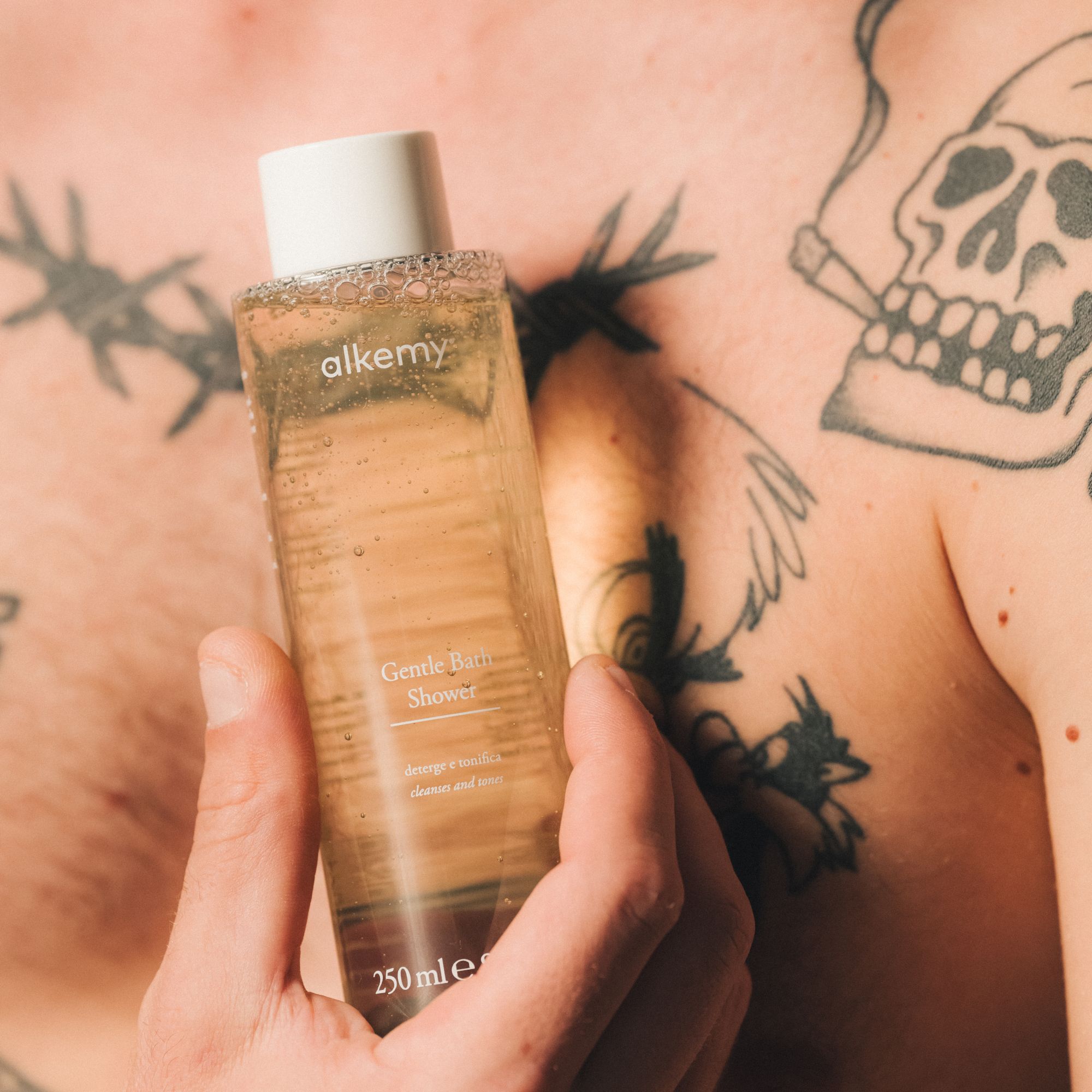Gel douche Alkemy dans une bouteille transparente, tenu par une main devant une peau tatouée.