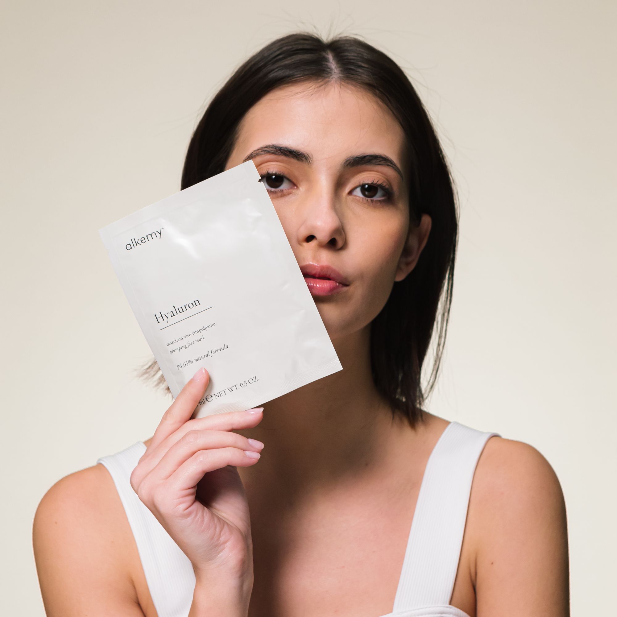 Une femme tient un sachet blanc de masque facial Alkemy Hyaluron devant son visage.