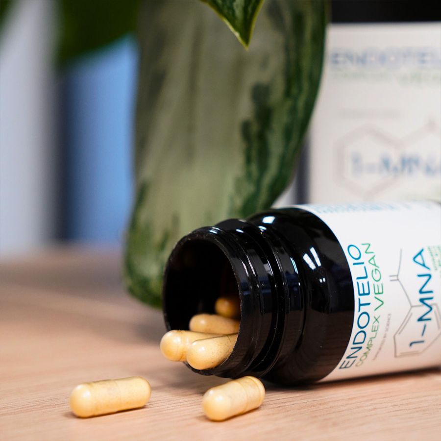 Bruine fles met capsules gevallen op een houten oppervlak. Opschrift: Endotelio Complex Vegan, 1-MNA. Enkele capsules ernaast.