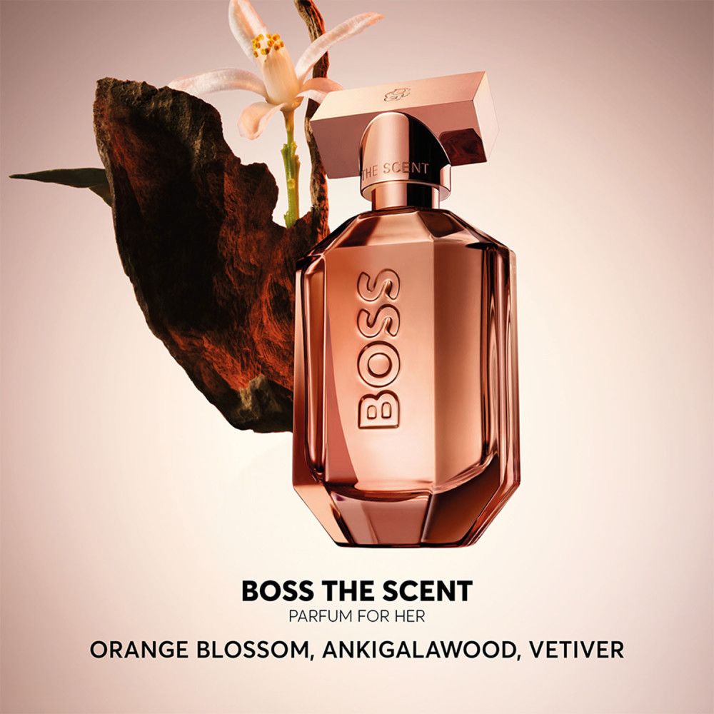 Parfumfles, koperkleurig, achthoekig. BOSS The Scent Parfum for Her. Bloemen en hout op de achtergrond.