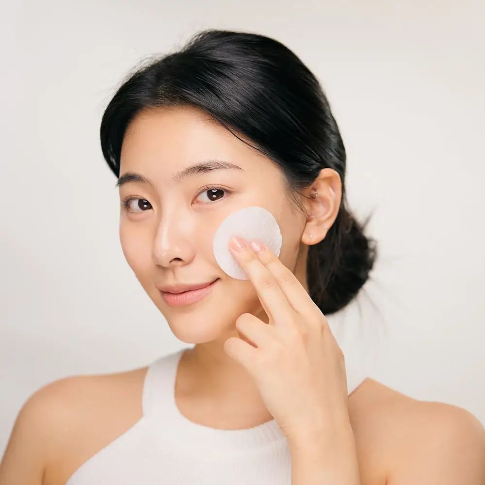 Jonge vrouw met een wattenschijfje op haar gezicht. Ze raakt haar huid aan met het schijfje. Lichte achtergrond.