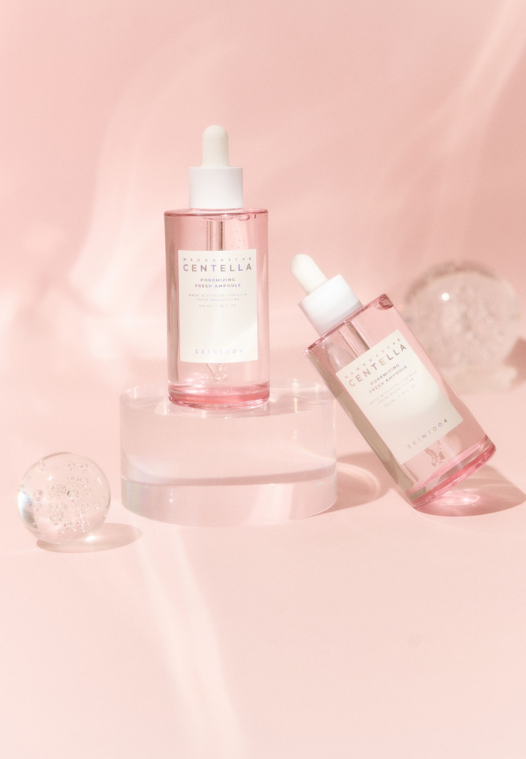 Deux flacons de sérum sur un piédestal. Fond rose. Billes de verre. Étiquette SKIN1004 visible.