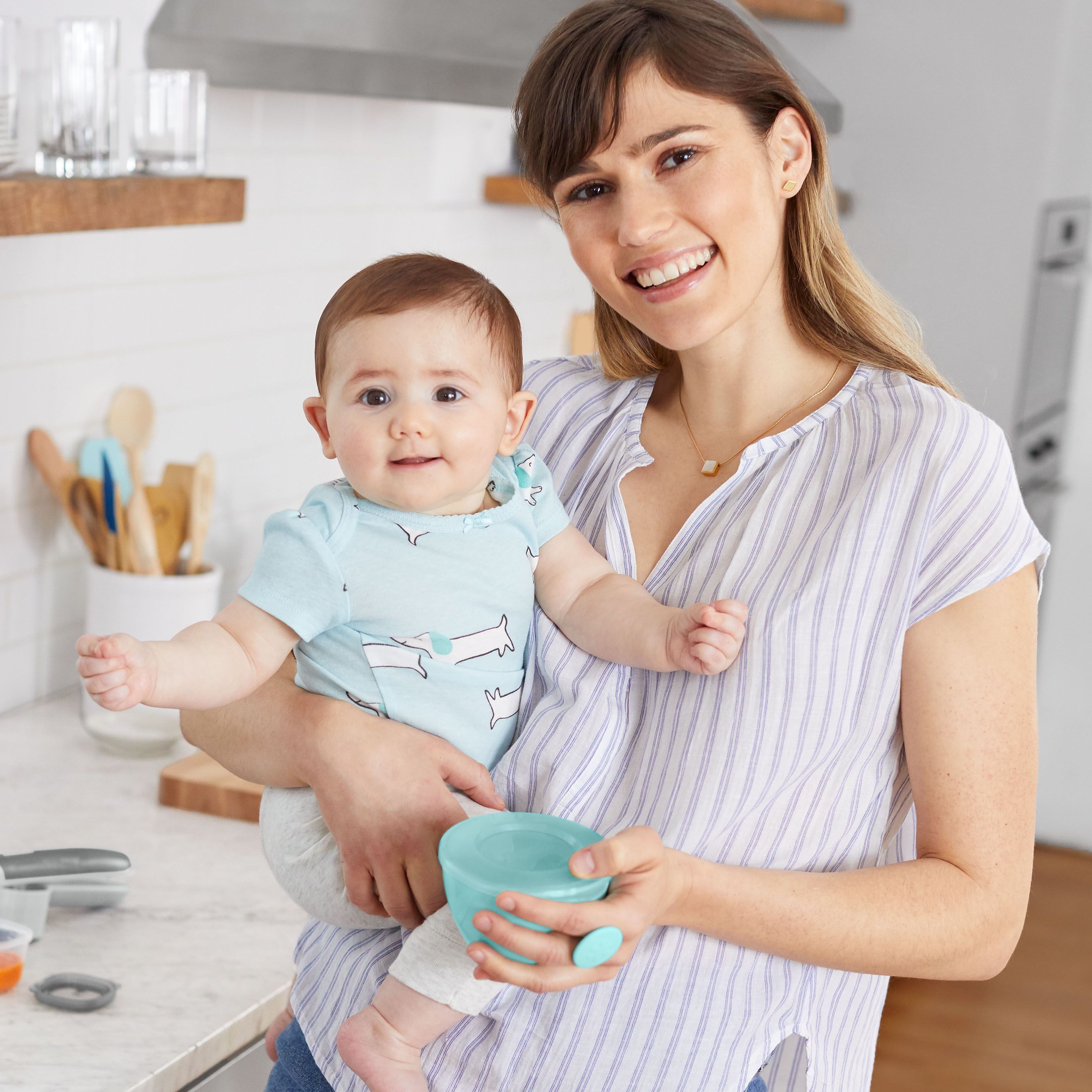 Een vrouw houdt een baby vast en een turquoise babyvoedingskom. De baby lacht.