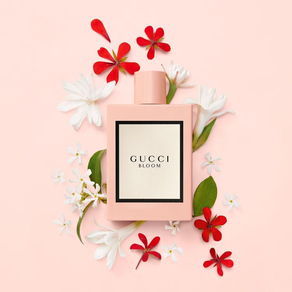 Roze fles omringd door witte en rode bloemen op roze achtergrond. Op de fles: Gucci Bloom, wit etiket met zwarte rand.
