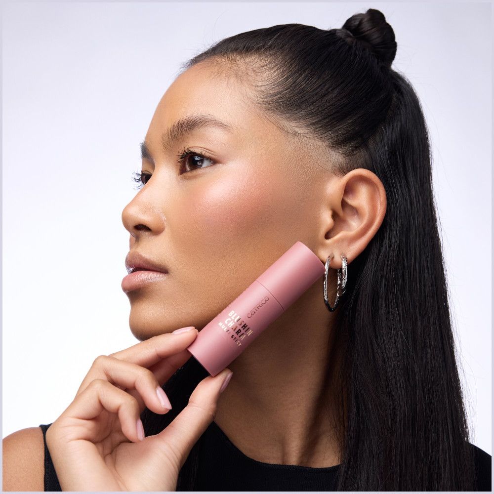 Femme tenant un Catrice Blush Stick. Fard à joues sur les joues. Créoles argentées.