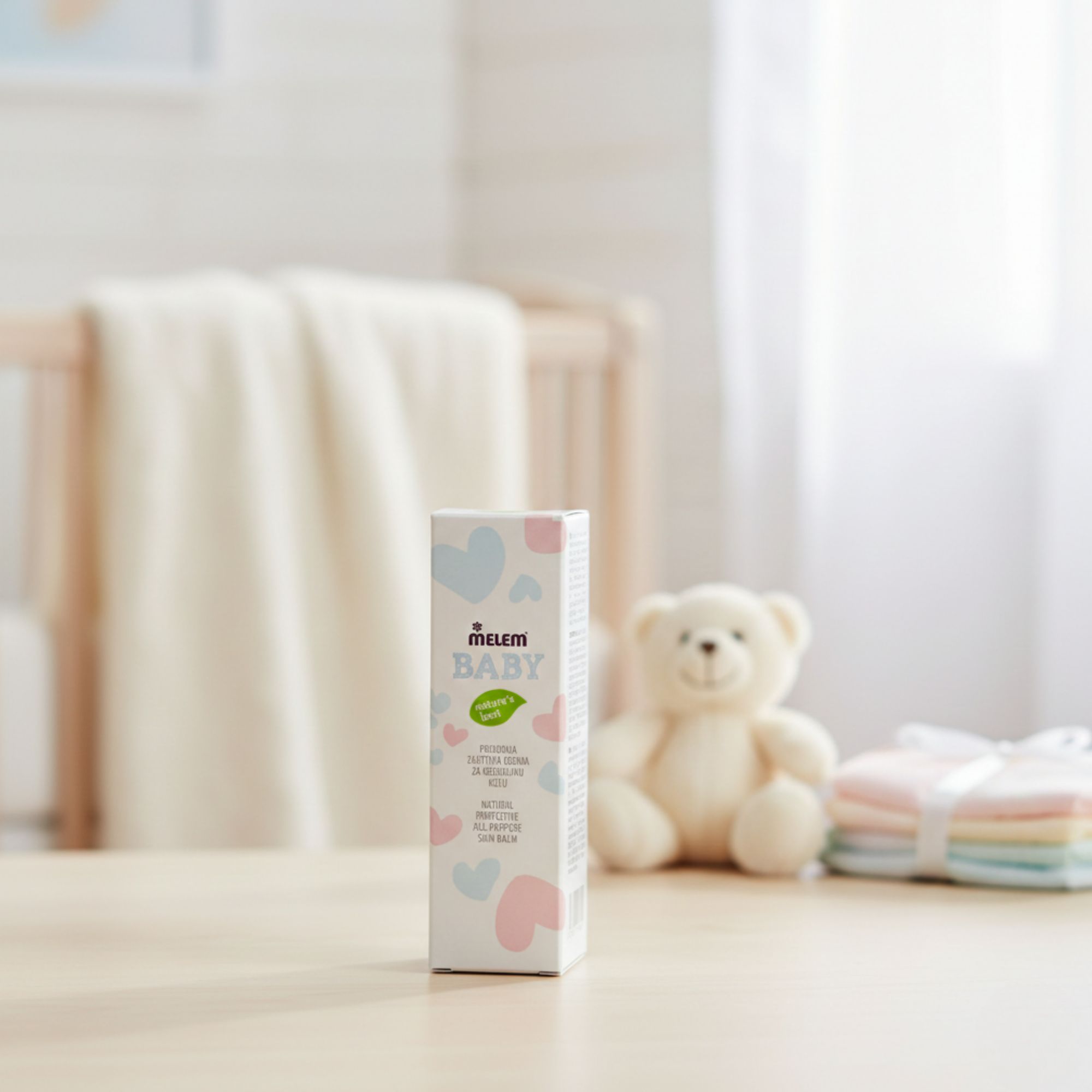 Boîte "Melem Baby" sur table. Texte : "Natural Protective All-Purpose Skin Balm". Arrière-plan : lit bébé, ours en peluche.