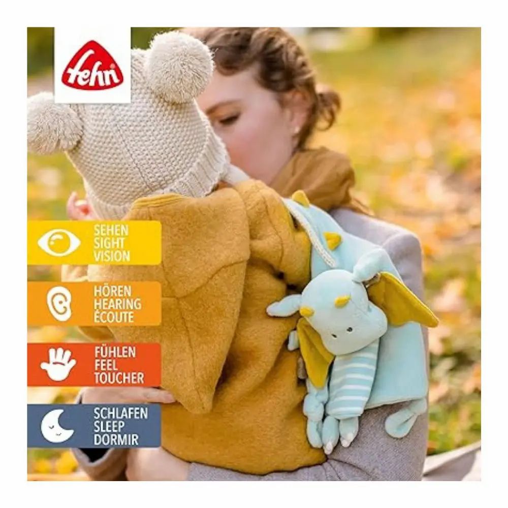 Baby met stoffen draak in de armen. Merk: Fehn. Symbolen voor zien, horen, voelen, slapen. Gele jas.