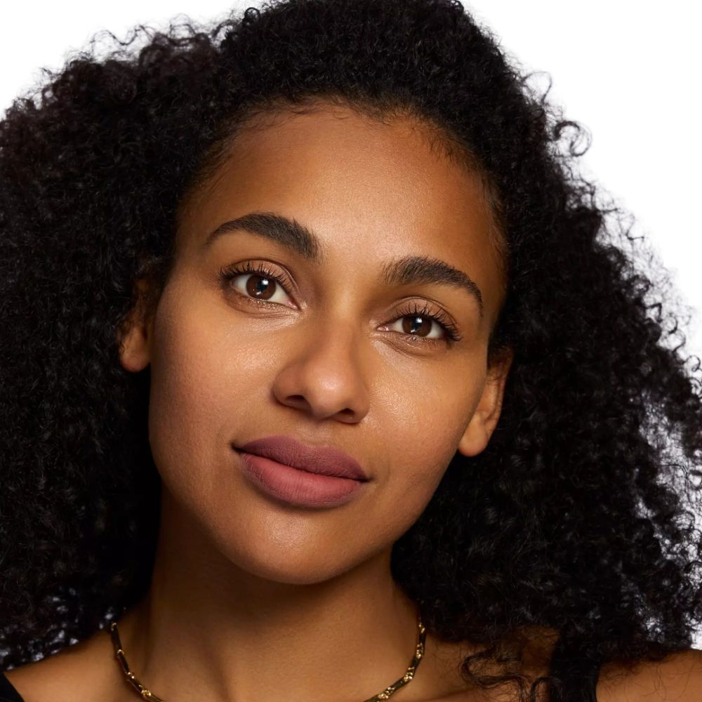Portrait d'une femme à la peau foncée et aux cheveux bouclés. Elle porte du rouge à lèvres mat.