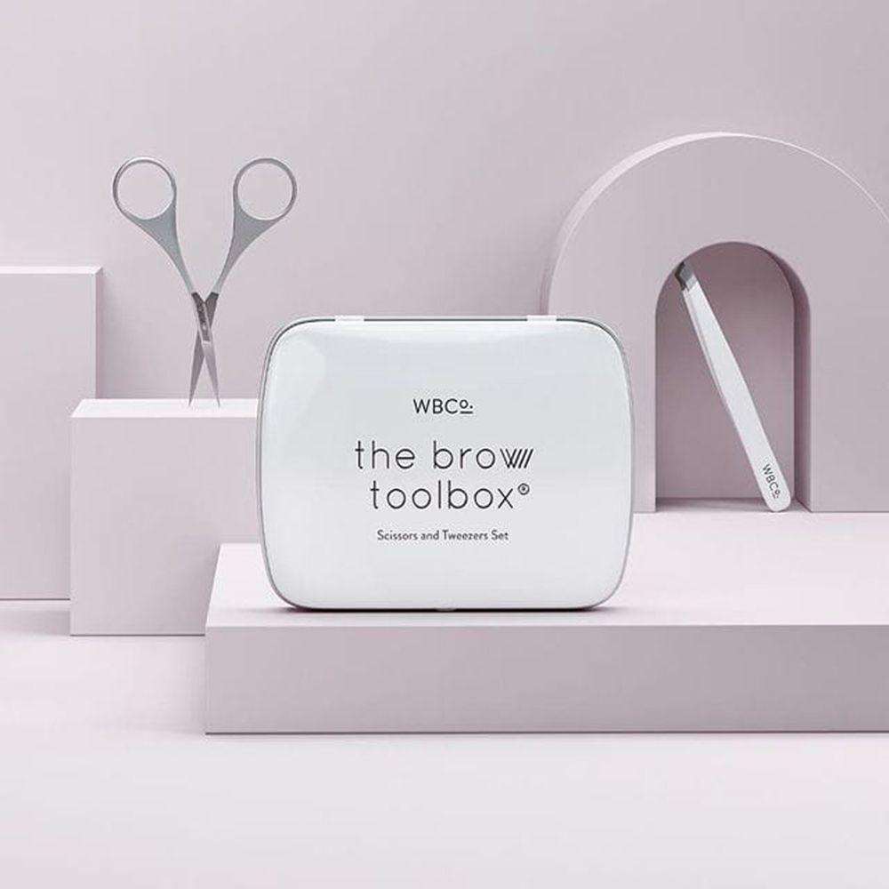 Trousse blanche avec inscription "the brow toolbox". Pince à épiler et ciseaux à côté. Marque : WBCo.
