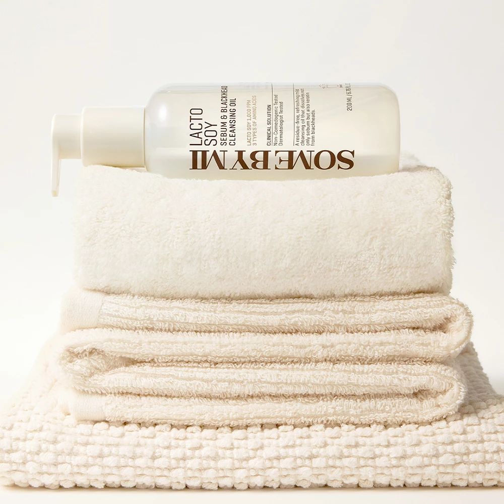 Flacon avec pompe, rempli de liquide, sur une pile de serviettes. Inscription : SOMEBYMI Lacto Soy Sebum & Blackhead Cleansing Oil.