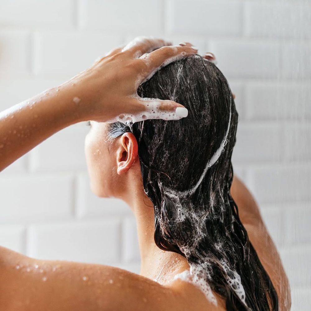 Personne se lave les cheveux sous la douche. Mousse sur les cheveux. Mains massant le cuir chevelu. Fond : carrelage blanc.