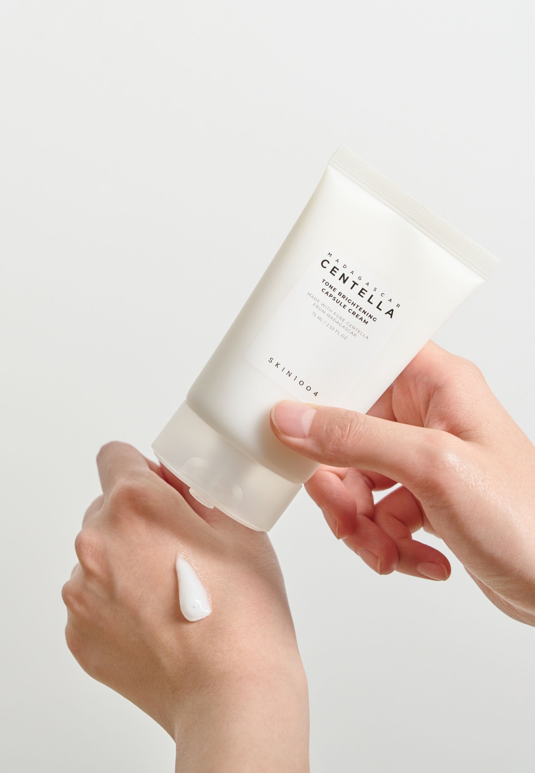 Handen houden een tube vast. Crème wordt op de hand aangebracht. Merk SKIN1004.