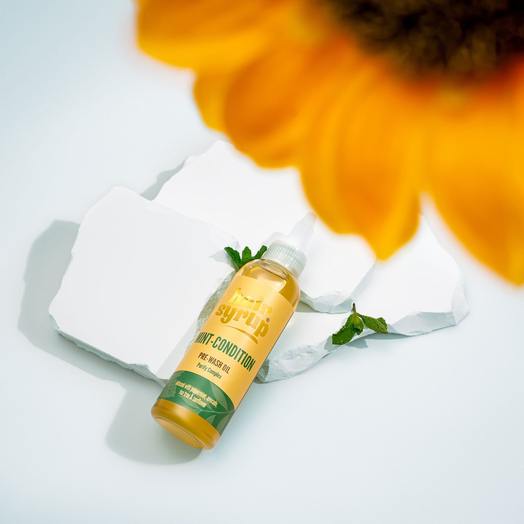 Gele fles "Hair Syrup Mint Condition" op witte stenen, munt en bloem op de achtergrond.
