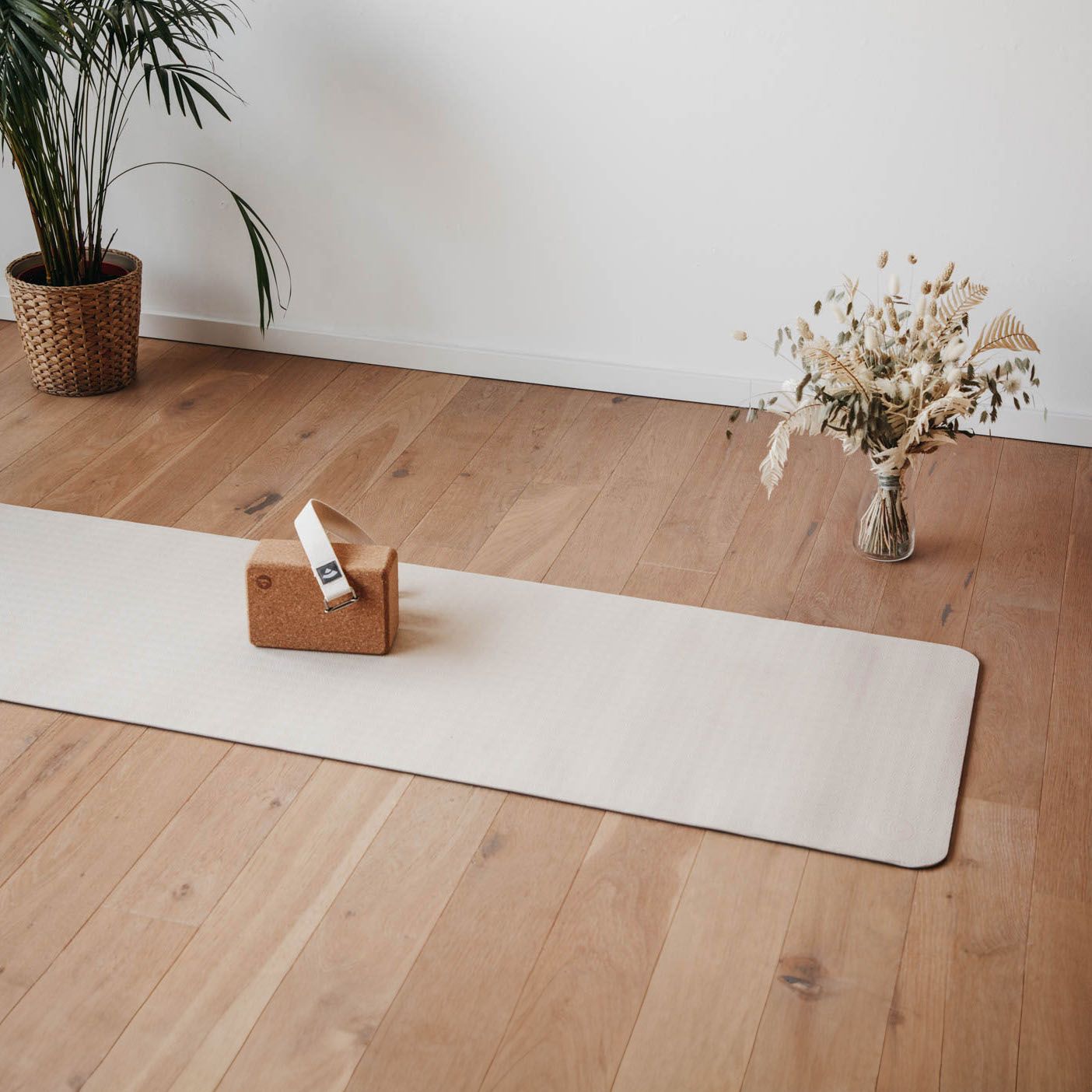Yogamat in lichte kleur. Op houten vloer. Met yogablok en tas. Plant en bloemen.