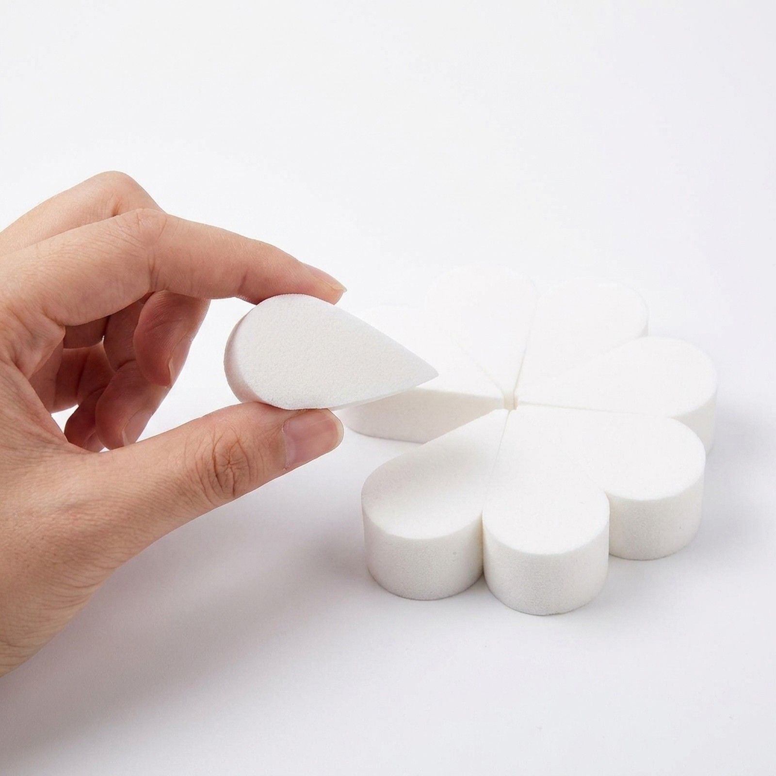 Een hand houdt een enkele witte, druppelvormige make-upspons vast. Op de achtergrond is een cluster van zes andere bloemvormige sponzen zichtbaar.