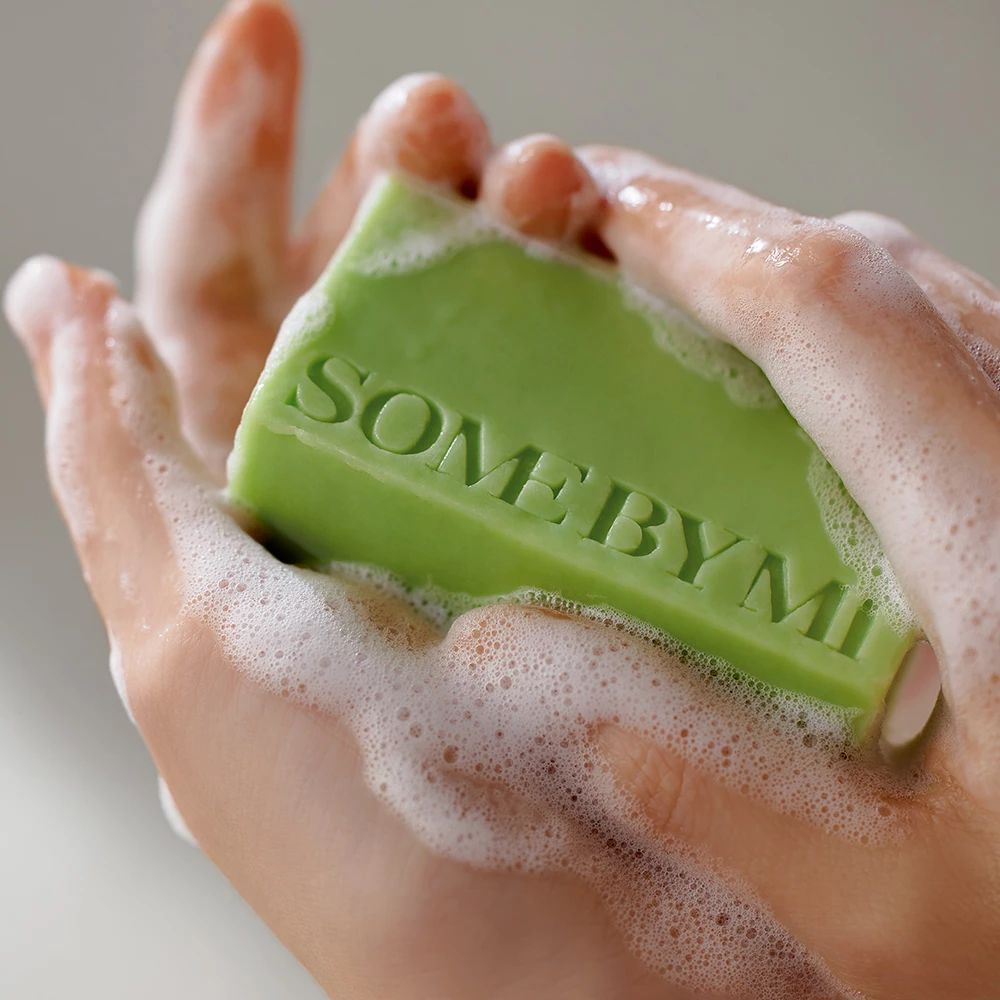 Pain de savon vert dans les mains, entouré de mousse. Le texte SOME BY MI est visible. Les mains sont couvertes de mousse savonneuse.