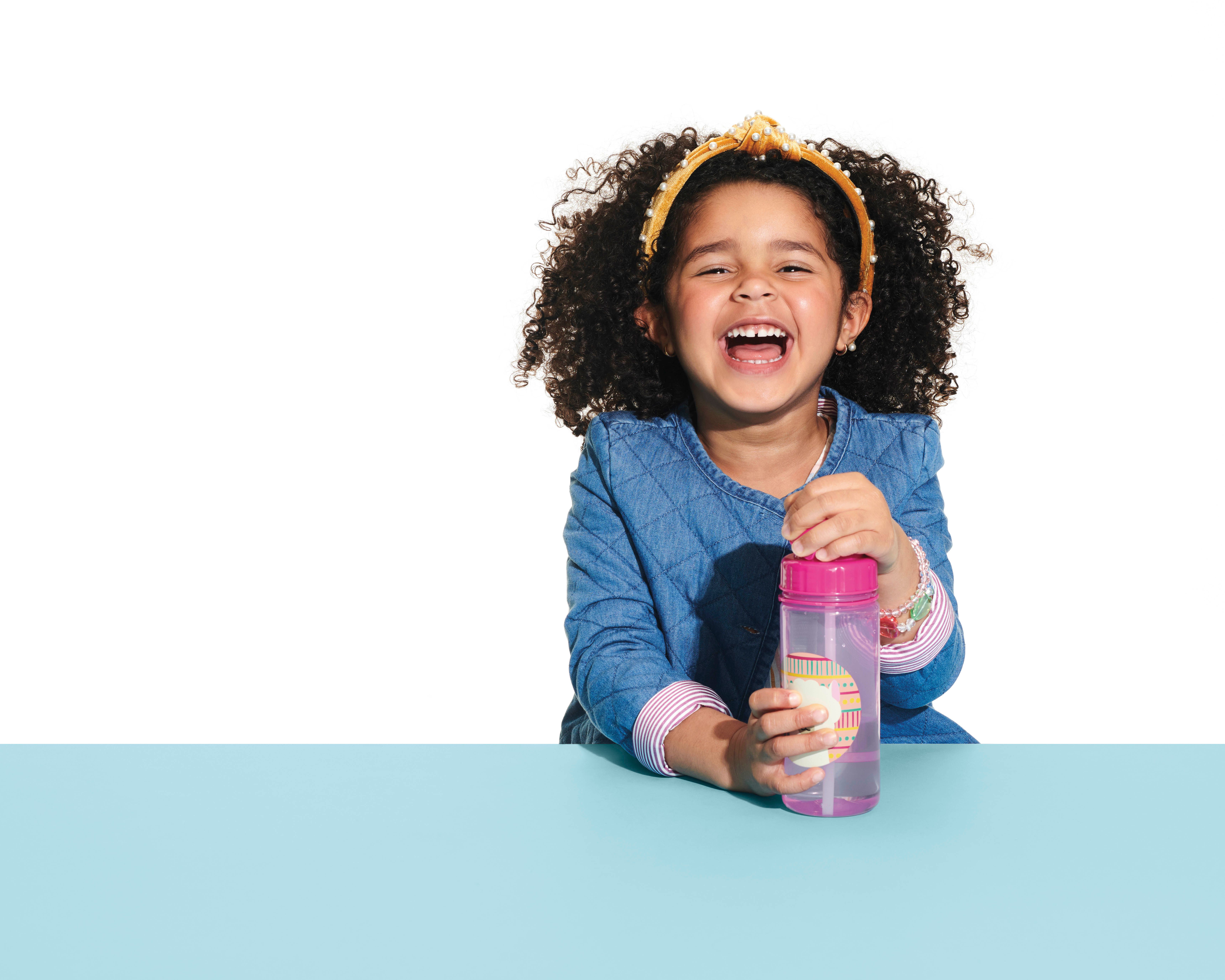 Lachend kind houdt roze drinkfles met lama-motief vast. Kind draagt spijkergoedhemd en haarband. Blauw tafelblad.