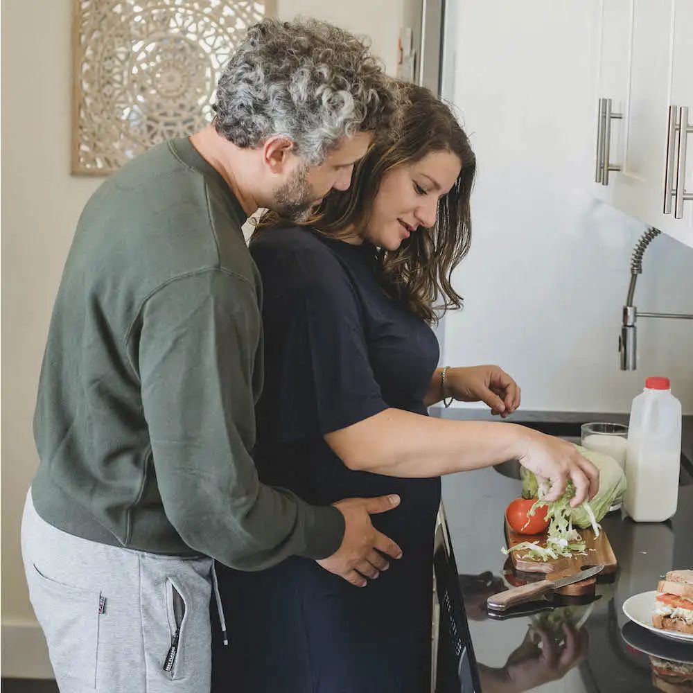 Een stel in de keuken. Vrouw snijdt groenten, man omhelst haar. Zwangere vrouw.