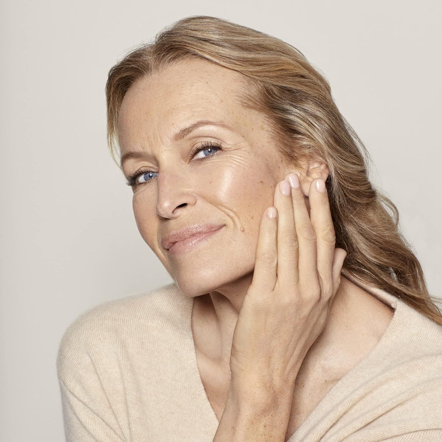 Femme touchant doucement sa peau du visage. Elle porte un pull beige. Son regard est dirigé vers la droite.