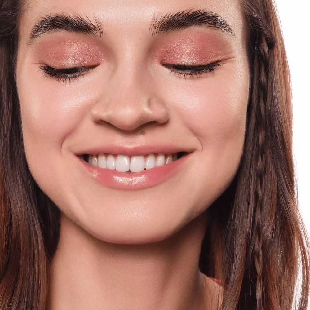 Une femme aux yeux fermés avec de l'ombre à paupières rose. Elle sourit.