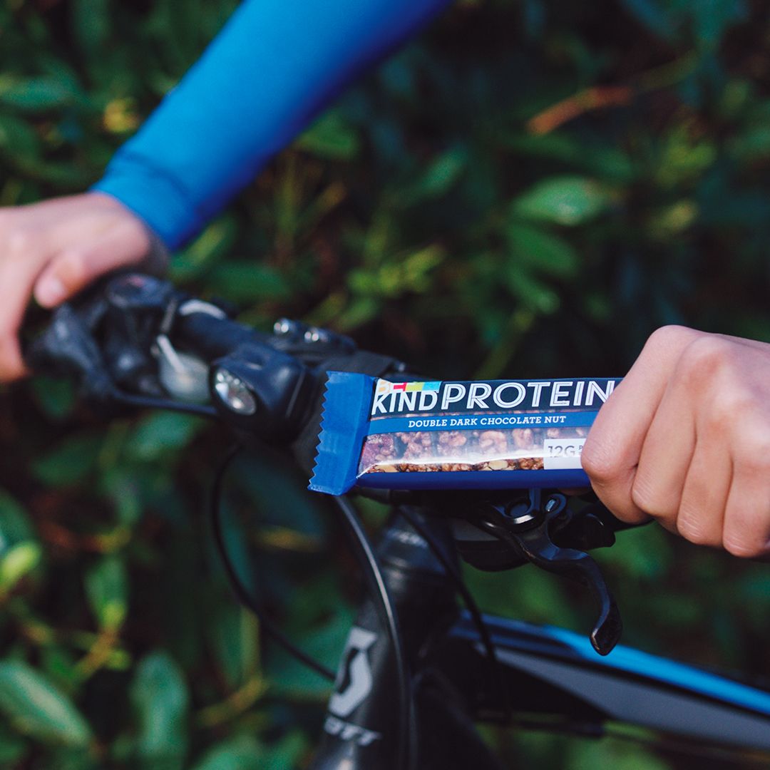 Persoon houdt een reep met pure chocolade en noten op een fietsstuur. Blauwe verpakking met logo en tekst.