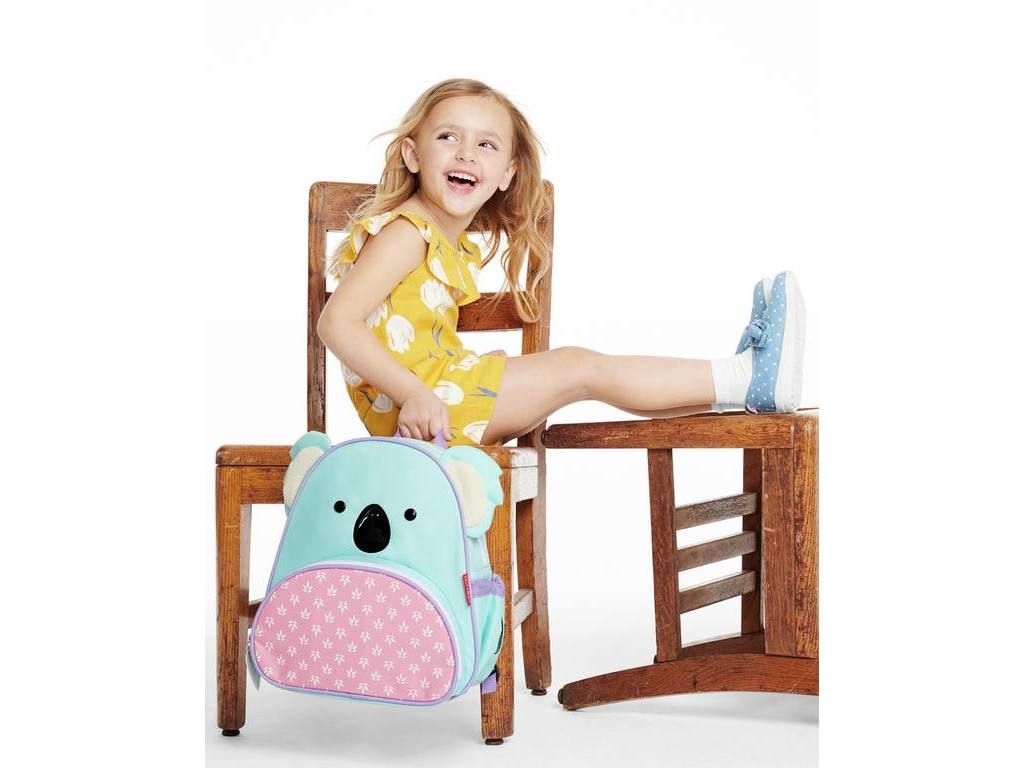 Kind zit op stoel met koala rugzak. Rugzak heeft oren, ogen en roze bodem. Merk Skip Hop.