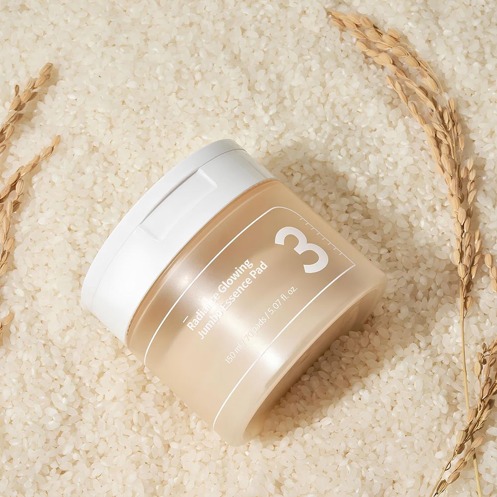 Pot beige avec couvercle blanc. Inscription : 3, Radiance Glowing Jumbo Essence Pad. Placé sur du riz et des épis de céréales.