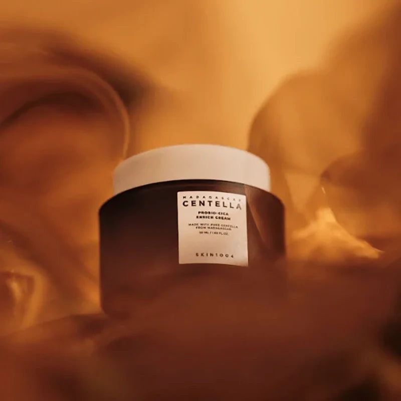 Pot de crème brun avec couvercle blanc, sur fond flou brun. Étiquette avec texte: MADAGASCAR CENTELLA, PROBIO-CICA ENRICH CREAM, SKIN1004.