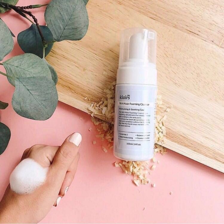 Klairs Rich Moist Foaming Cleanser in hand. Schuim op hand. Roze achtergrond met houten plank en planten.