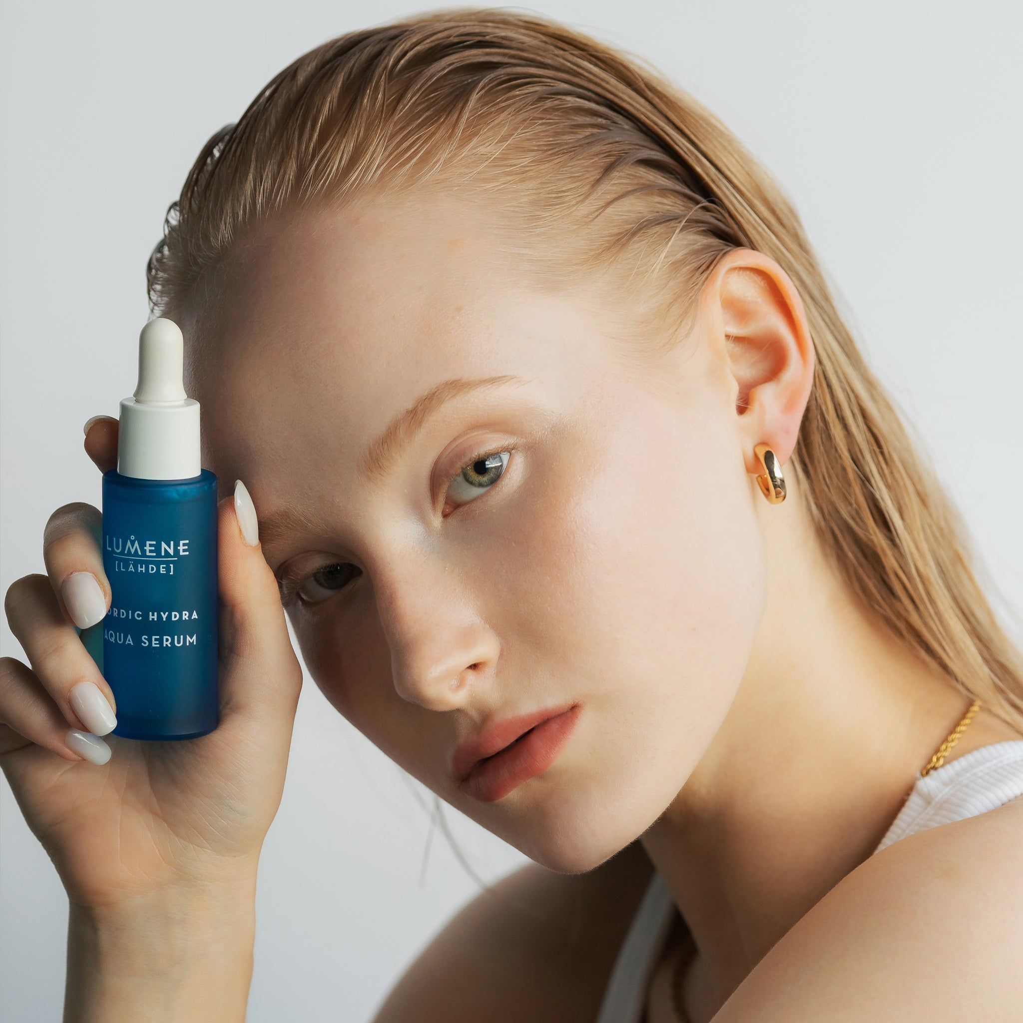Femme avec flacon de sérum. Inscription : Lumene [LAHDE] Nordic Hydra Aqua Serum. La femme porte des boucles d'oreilles dorées.