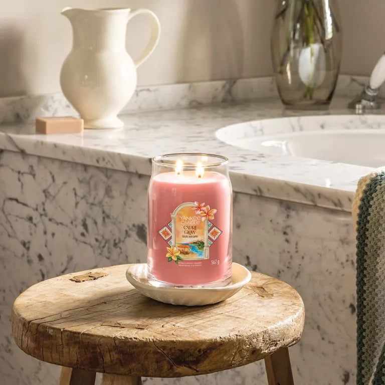 Bougie rose dans un verre, deux mèches, sur soucoupe. Inscription : Capri Glow. Sur tabouret. Arrière-plan salle de bain.