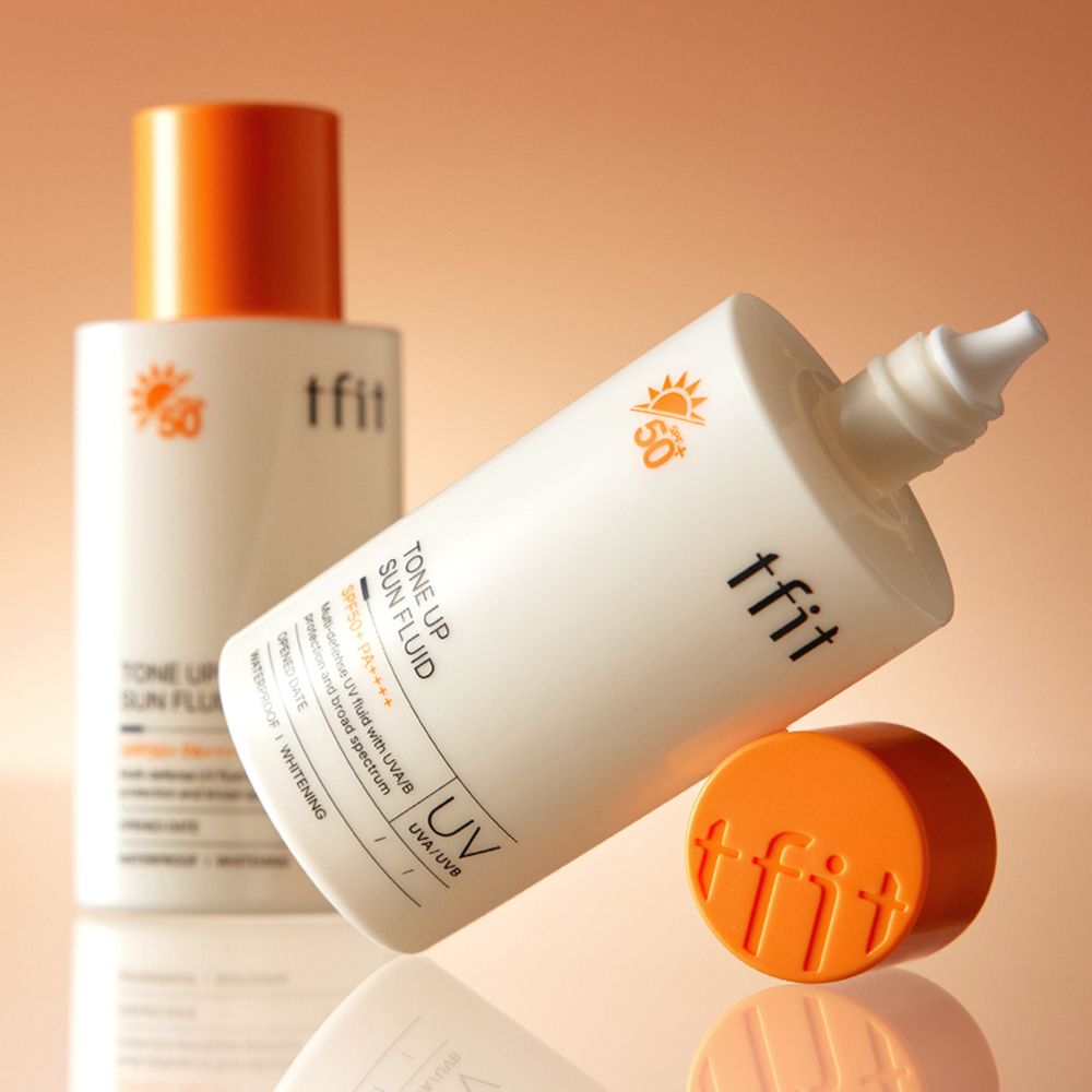Twee flessen, één liggend, één staand. Oranje doppen. Opschrift: TFIT, Tone Up Sun Fluid, SPF50+ PA++++. UV-beschermingssymbool.