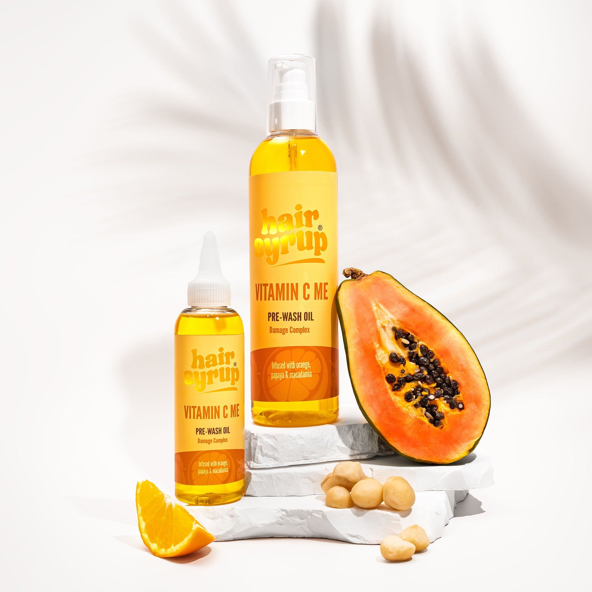 Twee flessen "Hair Syrup Vitamin C Me" en een halve papaja. Pre-Wash Oil. Op wit voetstuk.