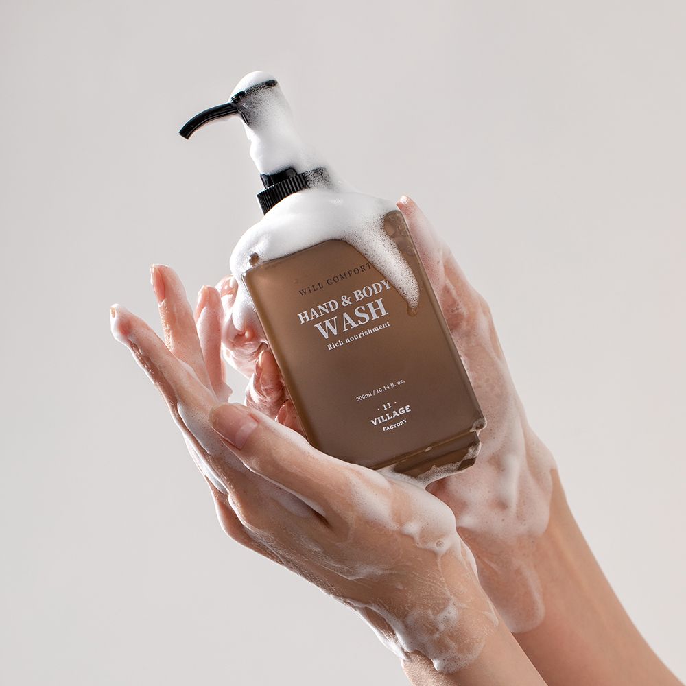 Handen houden een fles met schuim. Opschrift: WILL COMFORT HAND & BODY WASH. Merk: VILLAGE 11 FACTORY.