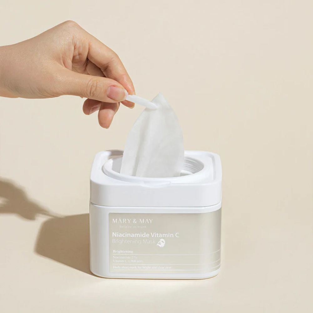 Hand die een masker uit een witte container met MARY & MAY Niacinamide Vitamin C Brightening Mask haalt.