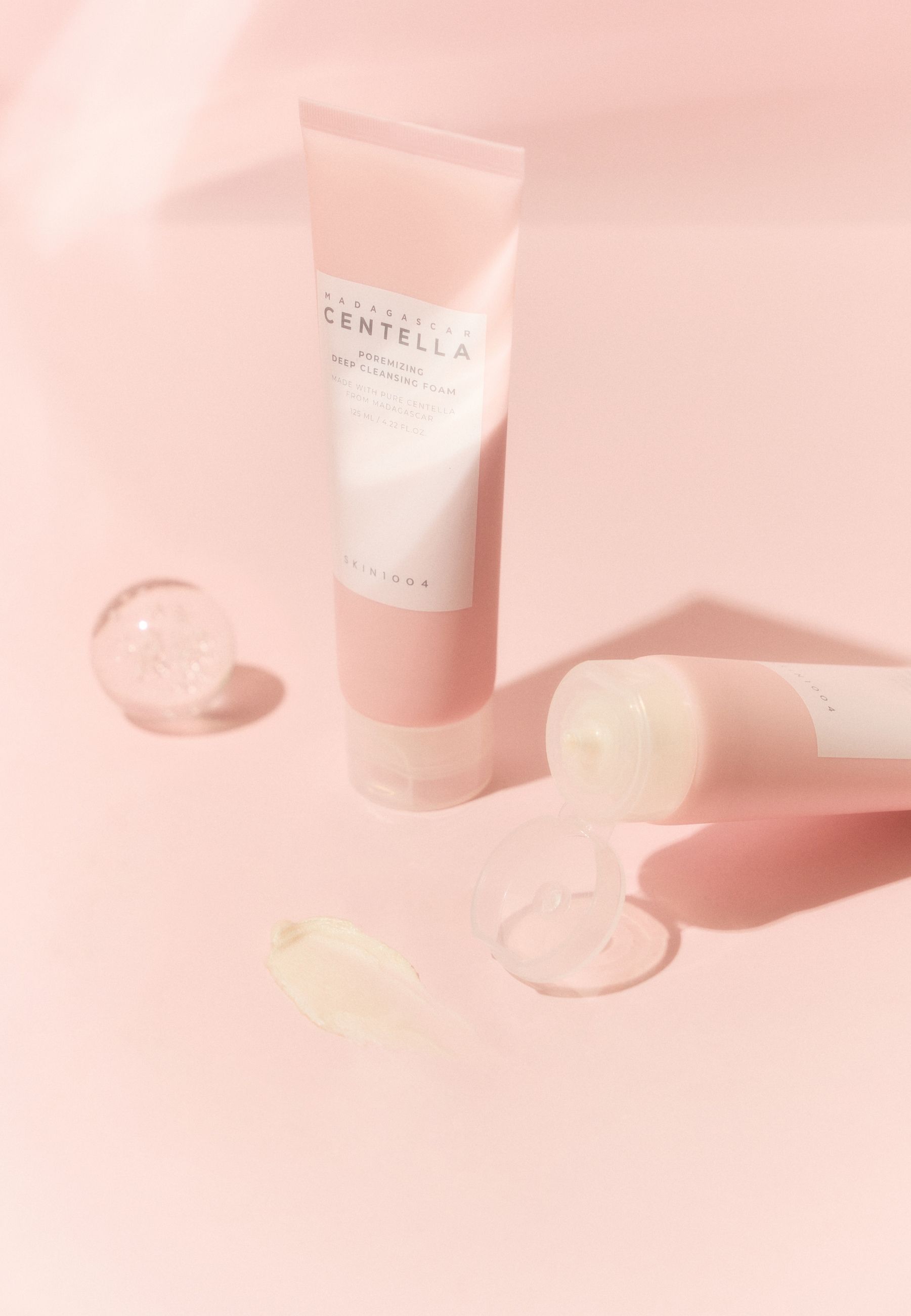 Deux tubes de mousse nettoyante rose, l'un ouvert, avec échantillon de mousse et sphère de verre.