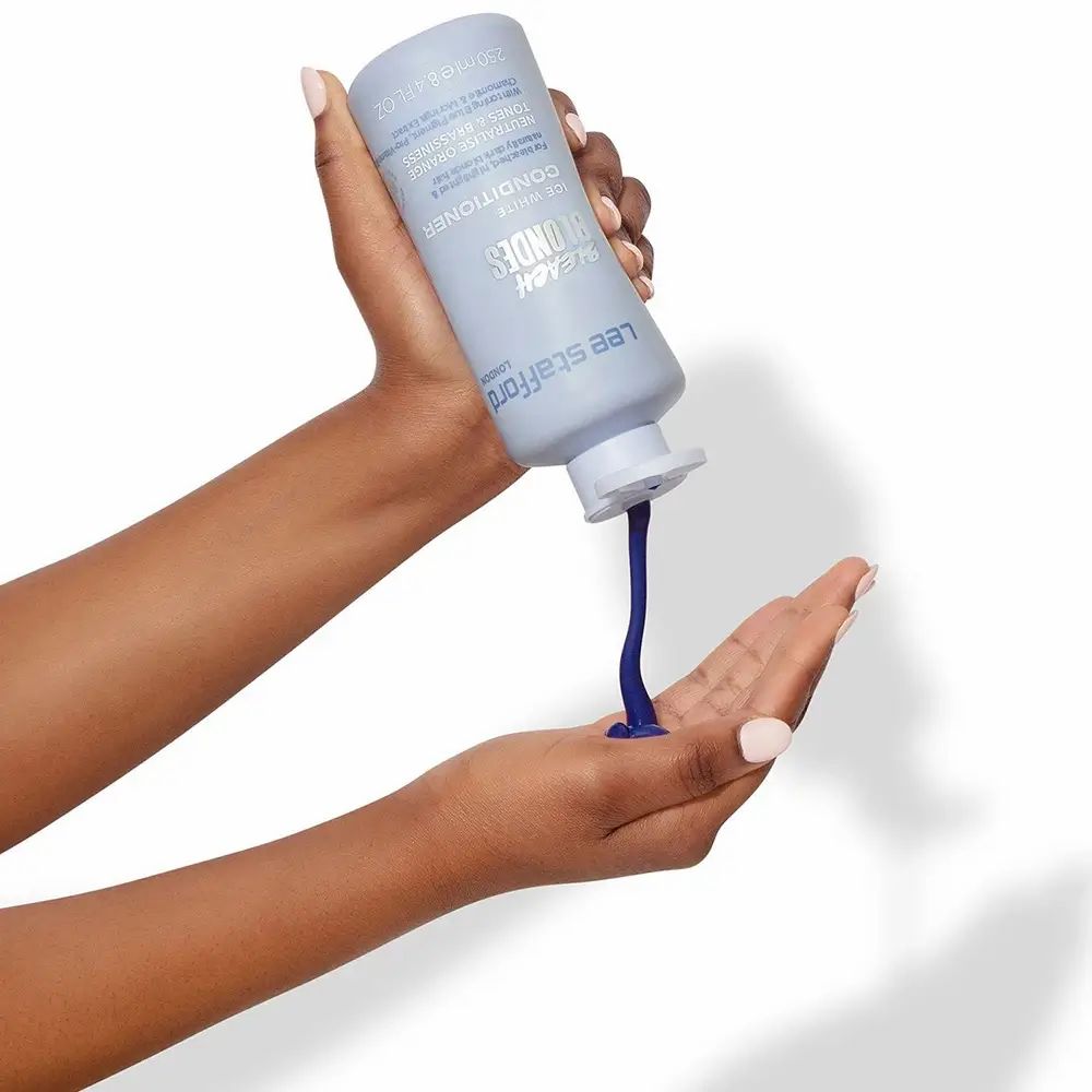 Handen houden een lichtblauwe fles vast. Product wordt in de handpalm gegoten. Product: LEE STAFFORD Bleach Blondes Conditioner.