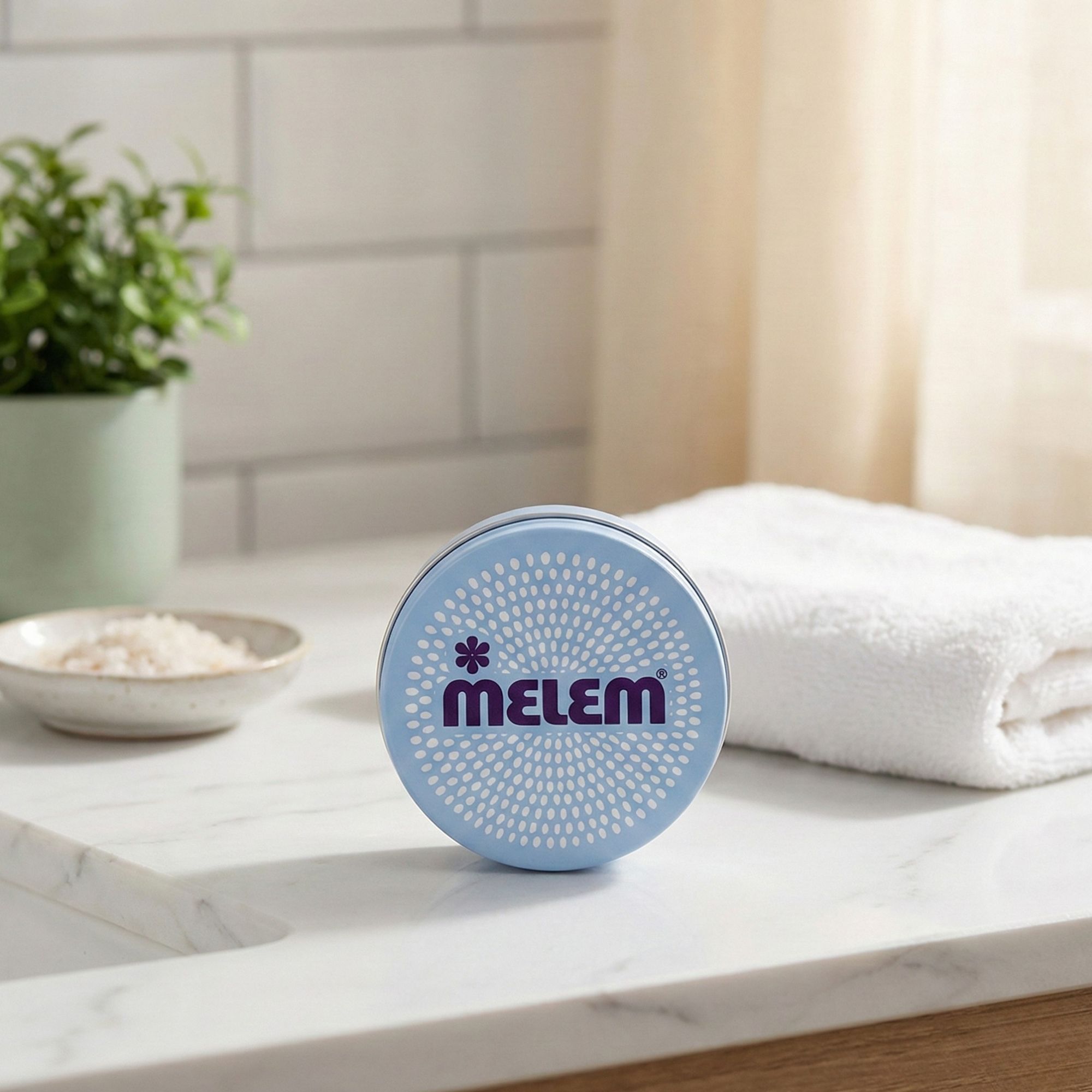 Lichtblauw, rond product met paarse tekst "MELEM". Op een wit oppervlak, naast een handdoek en een plant.