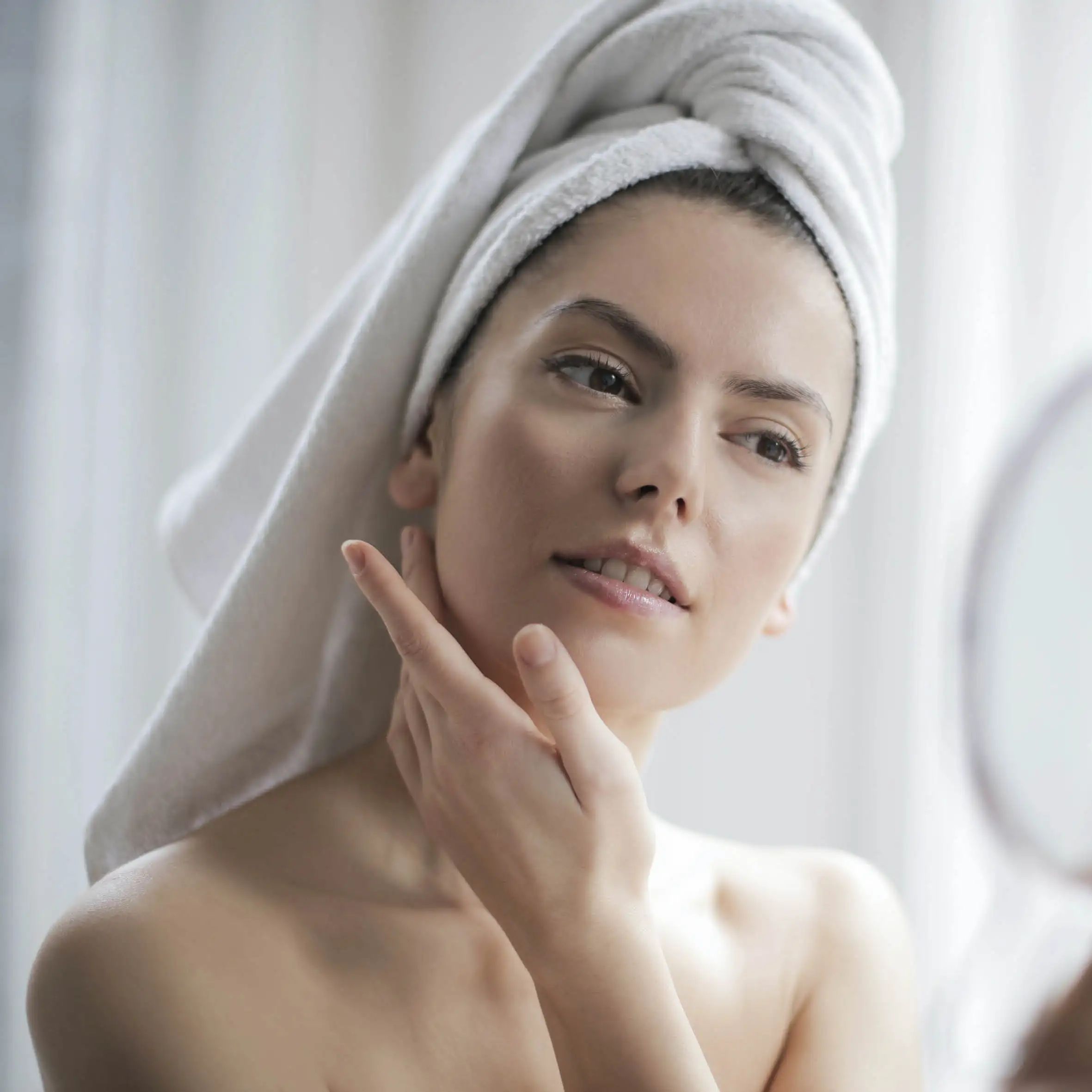 Femme, main sur le menton, regardant dans le miroir. Visage et épaules visibles. Serviette blanche sur la tête.