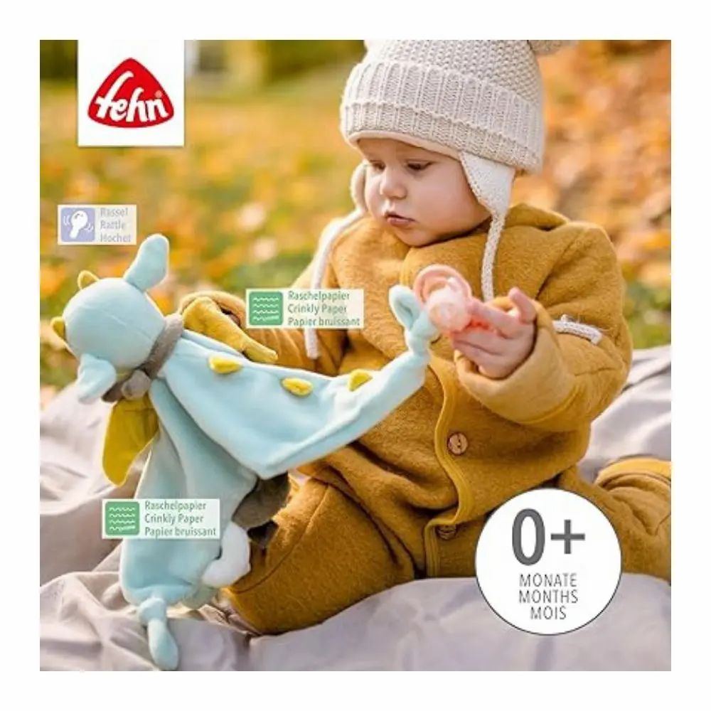 Baby houdt stoffen draak vast. Merk: Fehn. Tekst: "0+ MONATE MOIS MONTHS". Op de draak: knisperpapier.