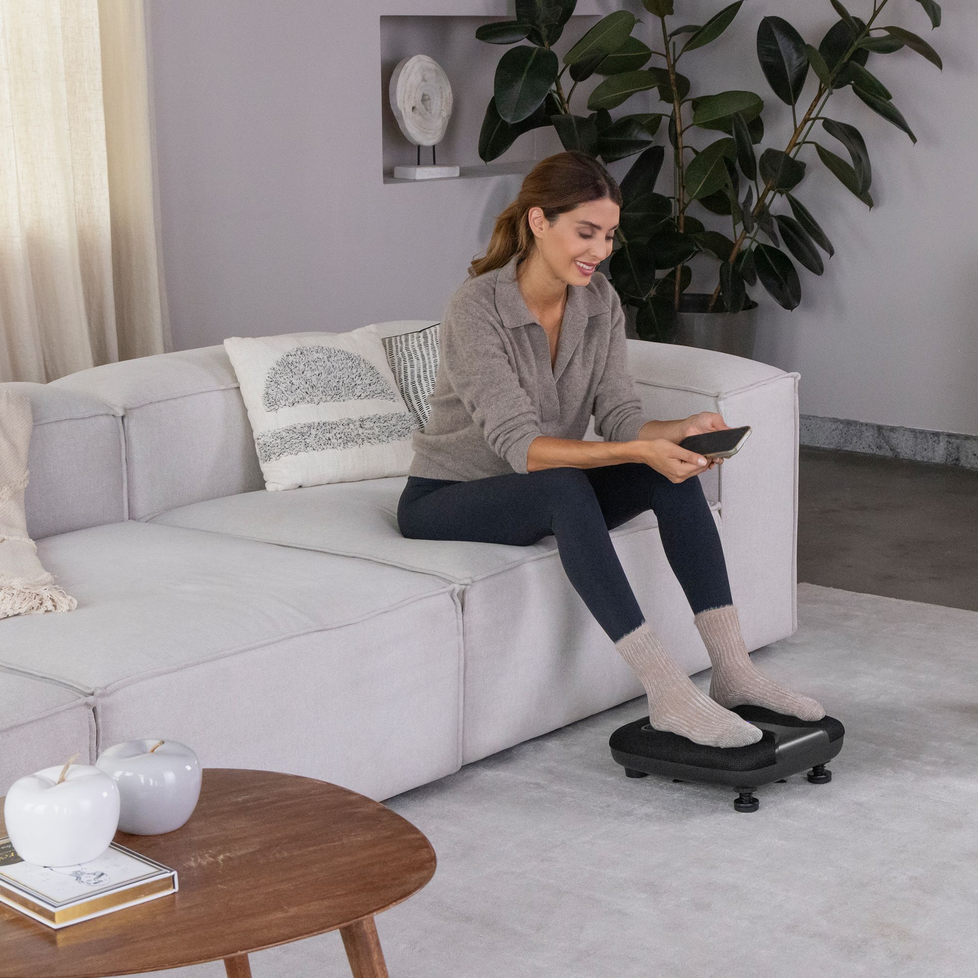 Vrouw zit op een bank en gebruikt een zwarte voetmassager. Voeten op het apparaat.