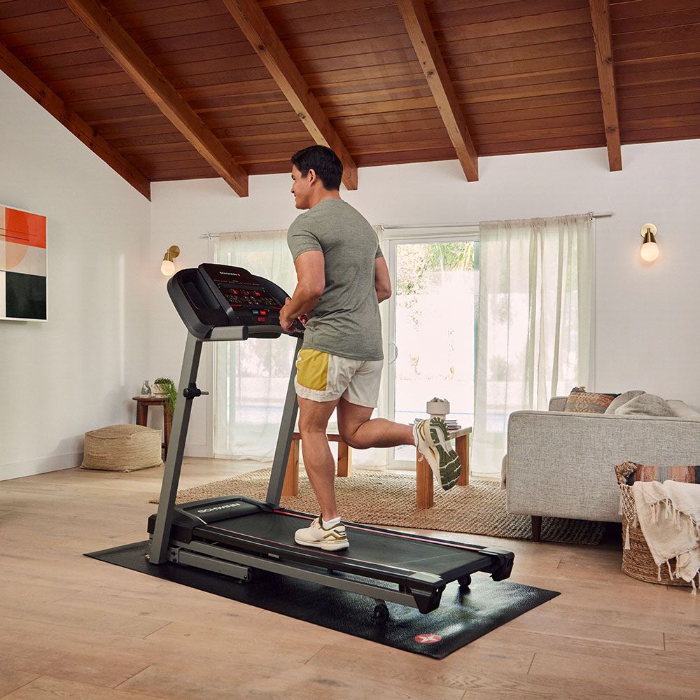 Homme courant sur un tapis de course. Surface noire, cadre gris. Console. Logo Schwinn. Dans un salon.