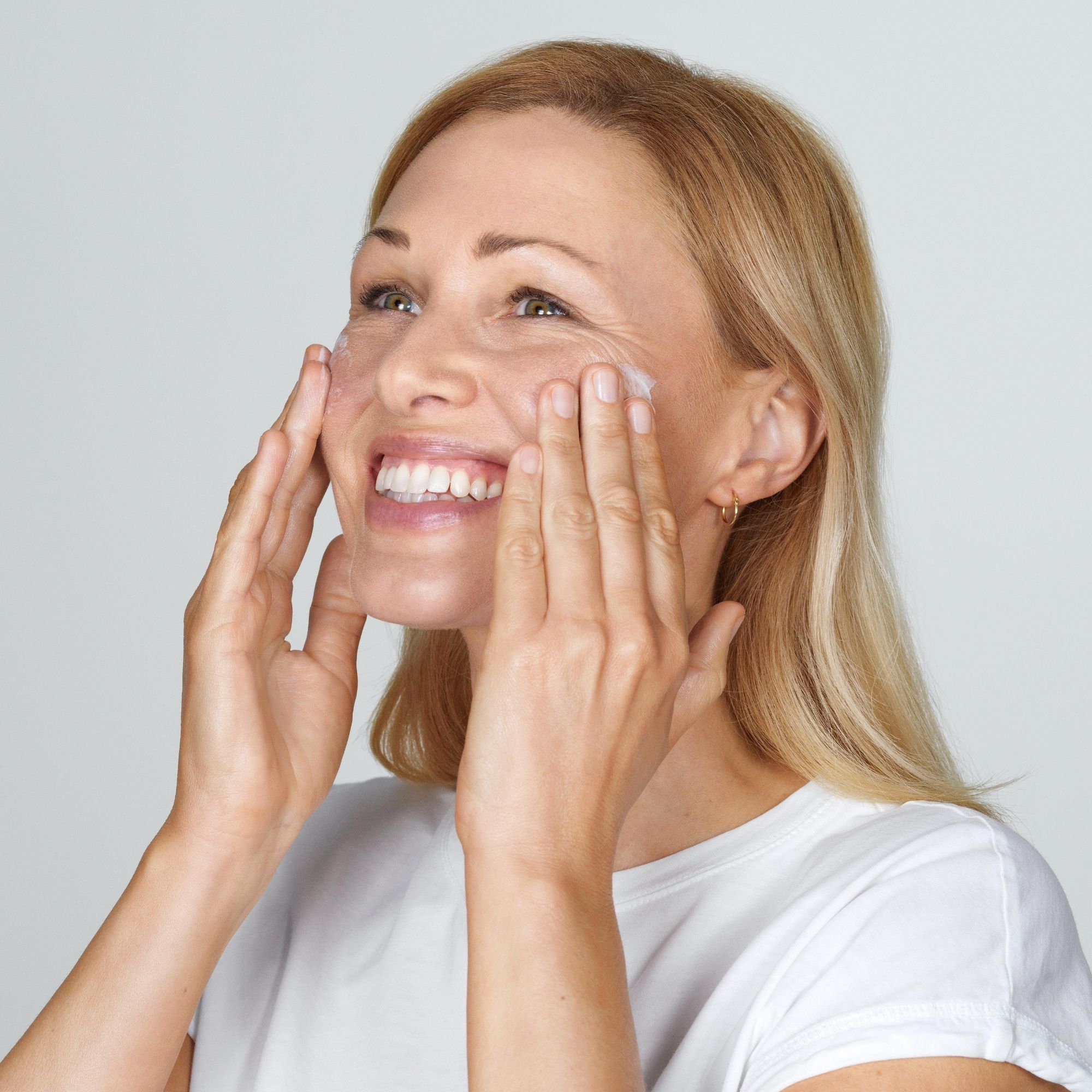 Femme appliquant de la crème sur son visage. Elle sourit et regarde vers le haut. Peau claire, cheveux blonds, t-shirt blanc.